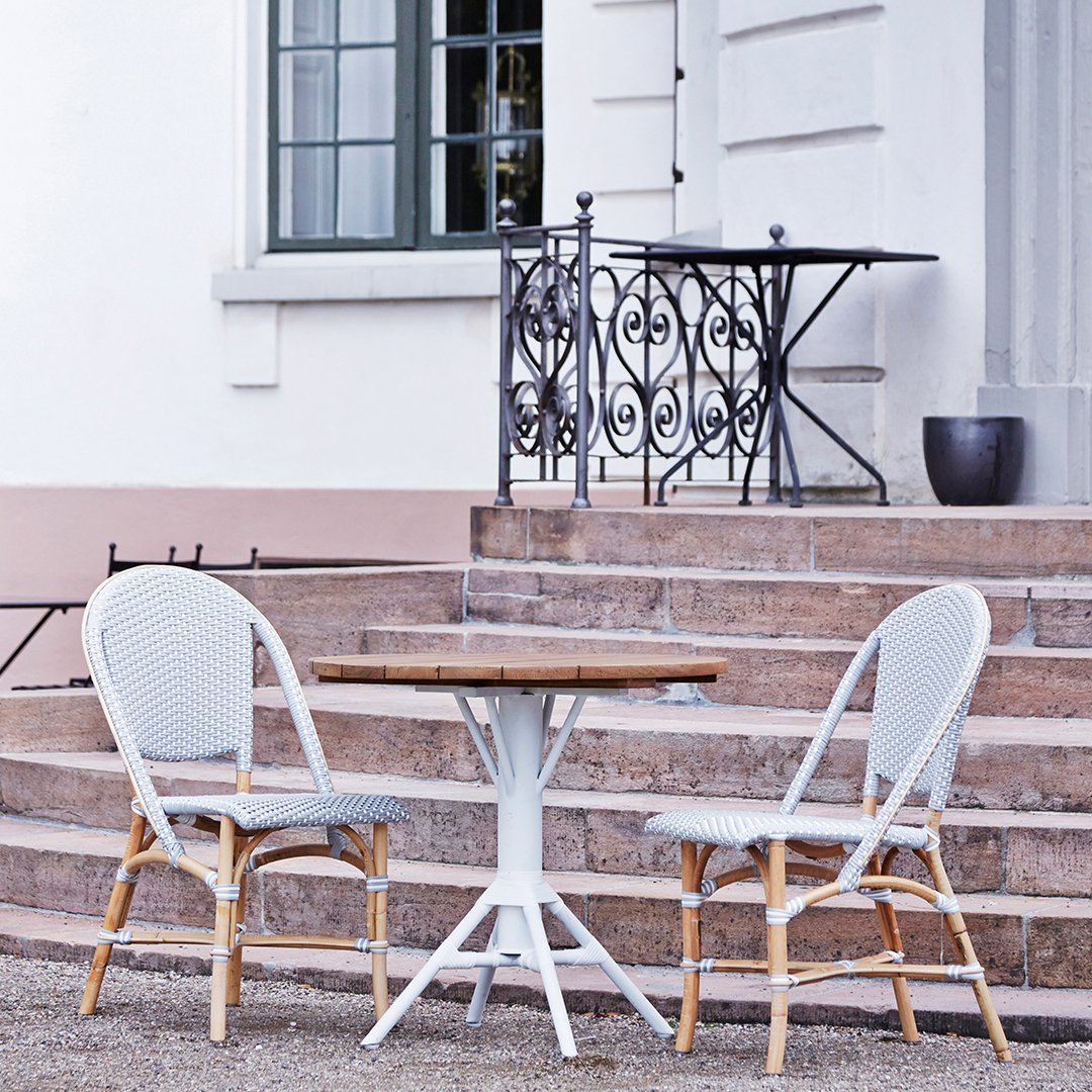 "White wicker chairs with a wood tabletop on a stone patio, featuring a soft gray backdrop and elegant black railing."