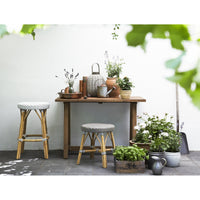"Natural wood table with gray stools, surrounded by green plants in clay and ceramic pots, creating a serene outdoor vibe."
