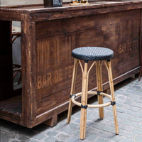 Bamboo bar stool with a woven black seat, set against a rustic wooden bar backdrop. Perfect for a stylish interior.