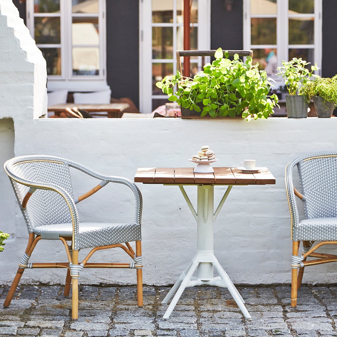 "Gray wicker chairs and a brown wooden table against a white wall, surrounded by green plants and natural stone flooring."