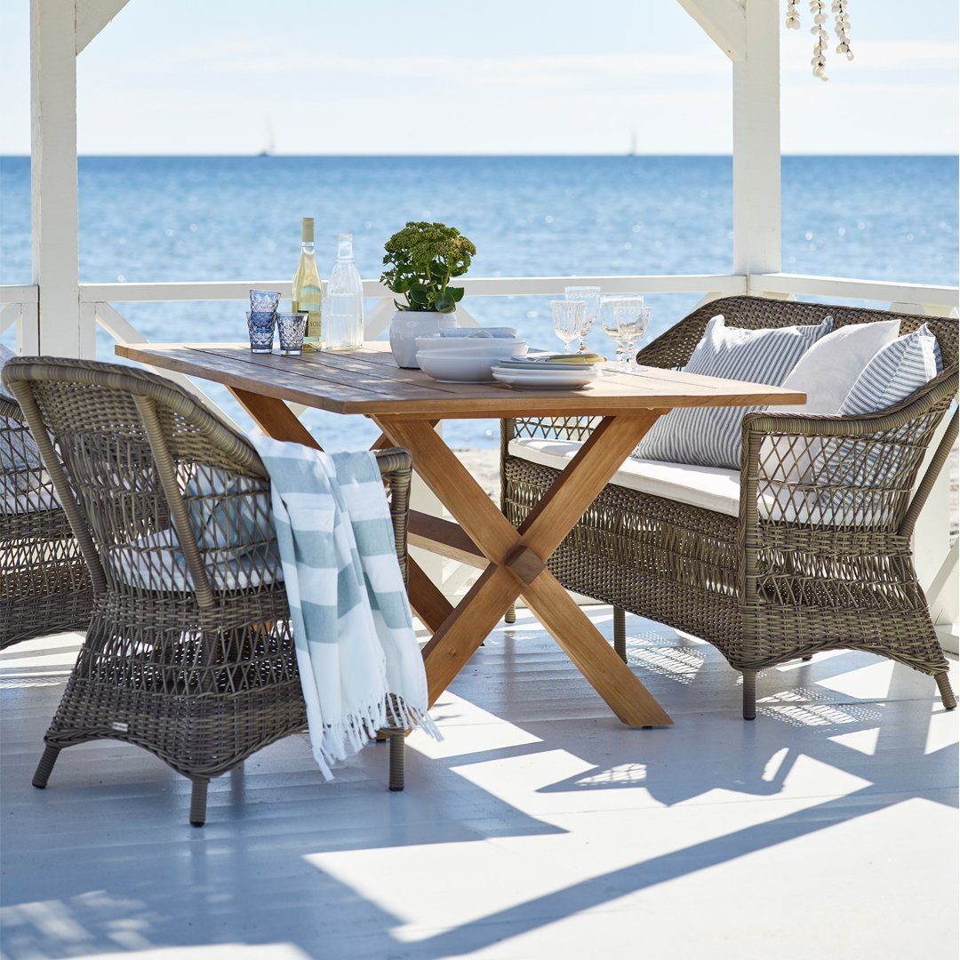 Coastal dining set featuring gray wicker chairs, blue-striped cushions, and a natural wood table, set against a beach backdrop.