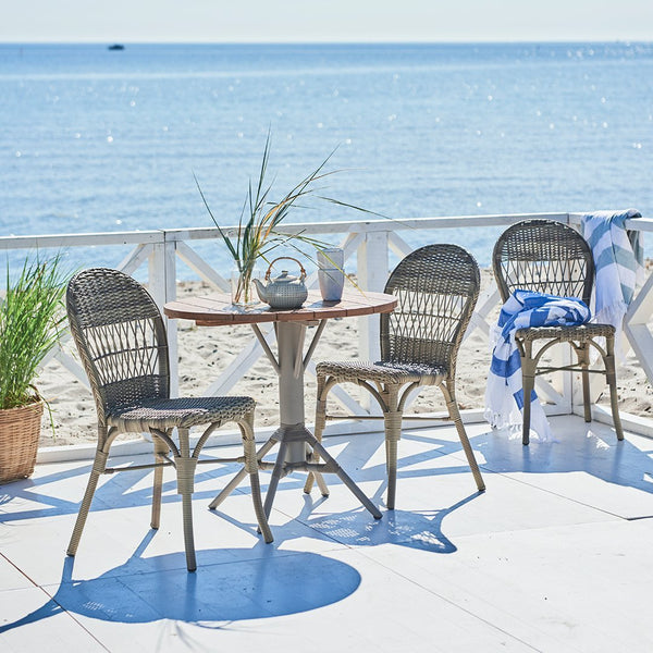 Gray woven chairs around a round wood table, set against a serene blue ocean backdrop, with green plants and white accents.
