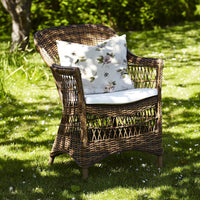 Woven brown wicker armchair with white cushion featuring floral patterns, set in a green garden.