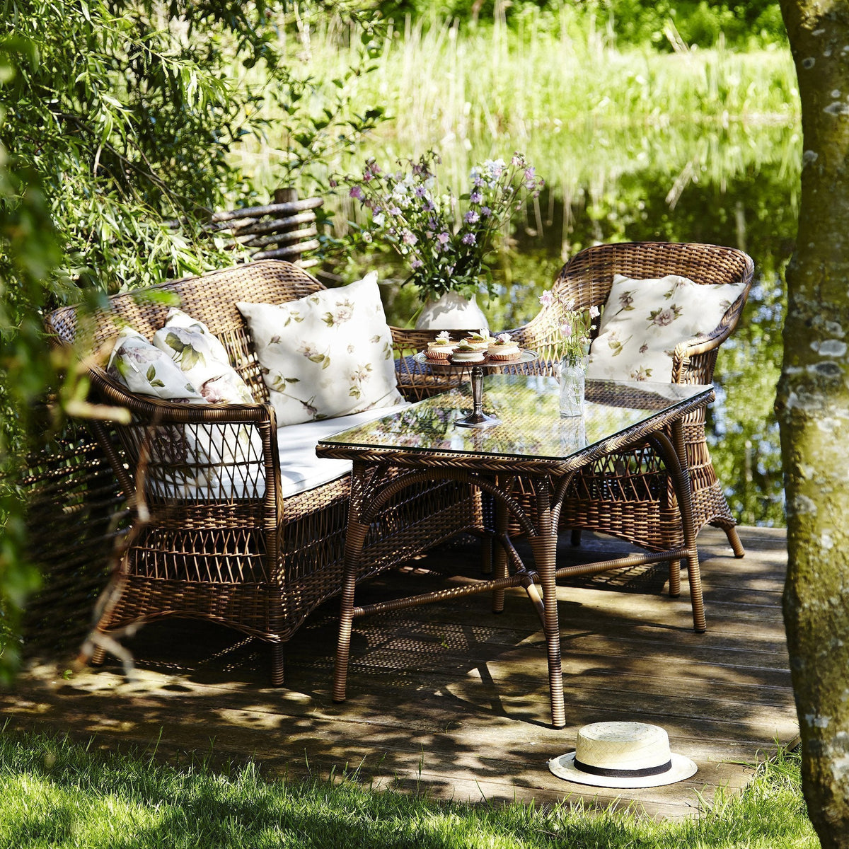 "Woven brown rattan outdoor furniture with floral-patterned cushions and a glass-top table, set by a serene lake."