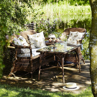 "Woven brown rattan outdoor furniture with floral-patterned cushions and a glass-top table, set by a serene lake."
