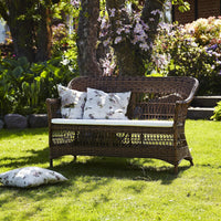 Brown wicker loveseat with floral-patterned cushions, set against a vibrant green lawn and blooming flowers.