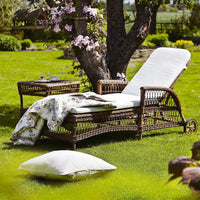 Wicker lounge chair with a white cushion, floral throw blanket, and a neutral pillow on lush green grass.