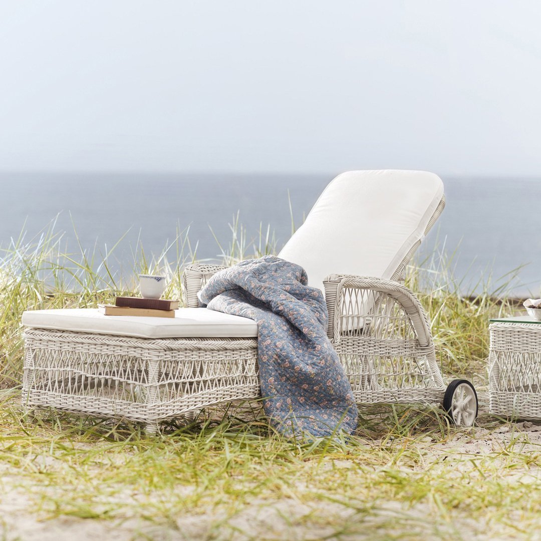 White wicker lounge chair with a light blue patterned blanket on sandy grass by the ocean.