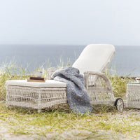 White wicker lounge chair with a light blue patterned blanket on sandy grass by the ocean.