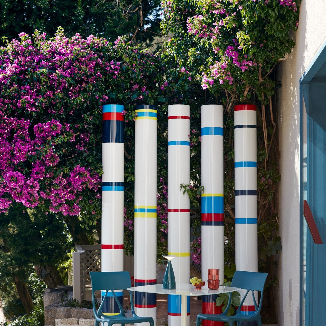 Colorful vertical poles with stripes in red, blue, yellow, and white, set against vibrant pink flowering plants.