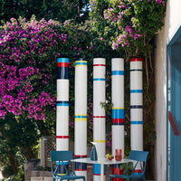 Colorful vertical poles with stripes in red, blue, yellow, and white, set against vibrant pink flowering plants.