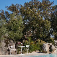 White outdoor chairs and table set against lush green foliage and natural stone, creating a serene poolside vibe.