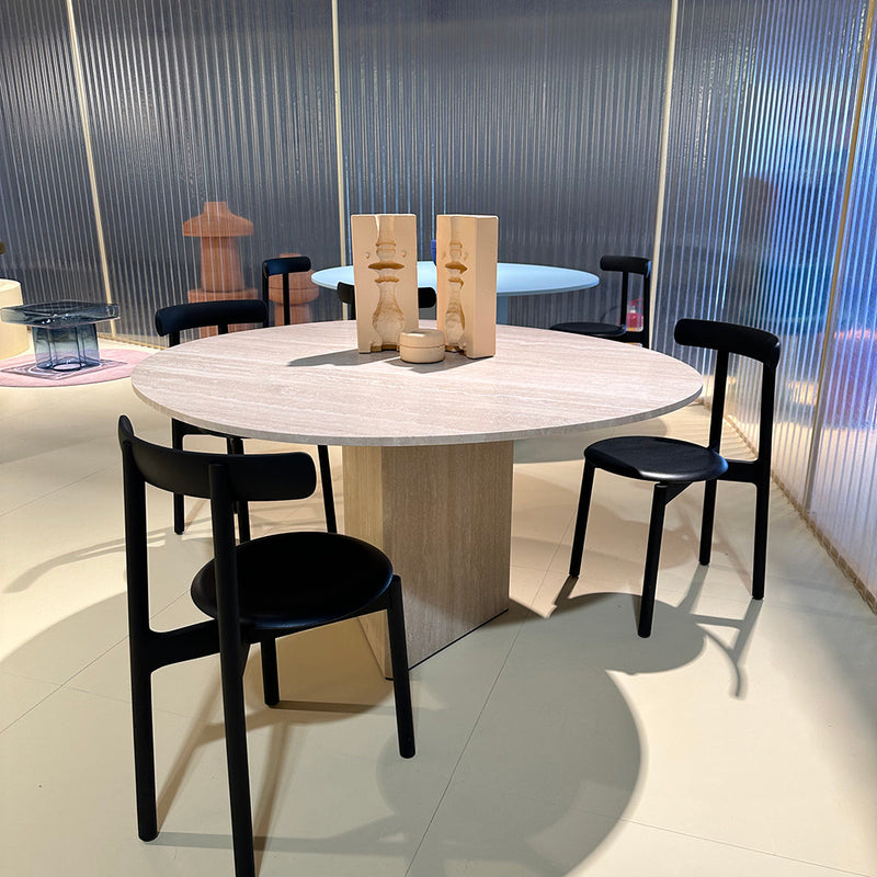 Round wooden dining table with a light finish, surrounded by minimalist black chairs and a soft, neutral background.