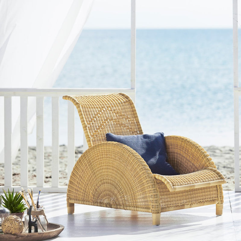 Natural woven rattan chair with a light tan finish and a deep blue cushion, set against a serene beach backdrop.