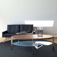 Modern oval coffee table with a wood finish, glass top, and silver legs; paired with a black sofa on a textured dark rug.