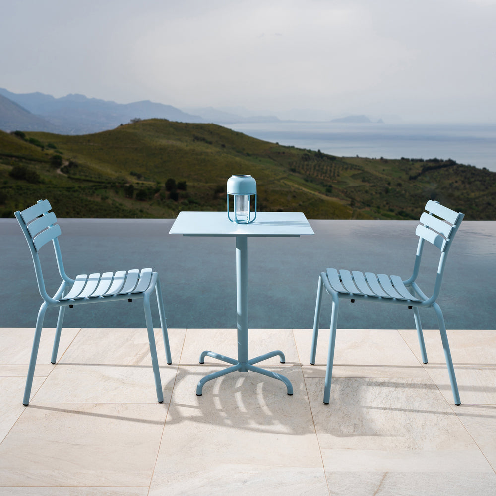 Sky blue outdoor table and chairs with sleek slatted design, set against a scenic backdrop of mountains and water.