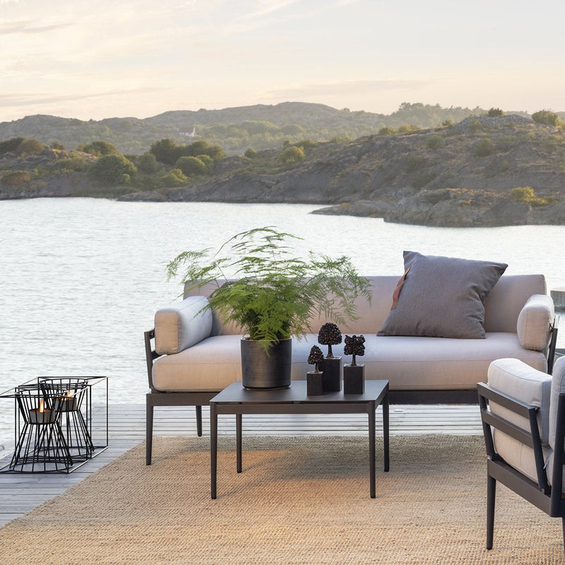 "Beige outdoor sofa with dark metal accents, paired with a black table and green plant, set against a serene water backdrop."