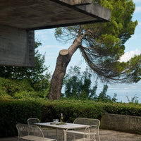 Outdoor dining set with white metal chairs and table, surrounded by lush green foliage and a clear blue sky backdrop.