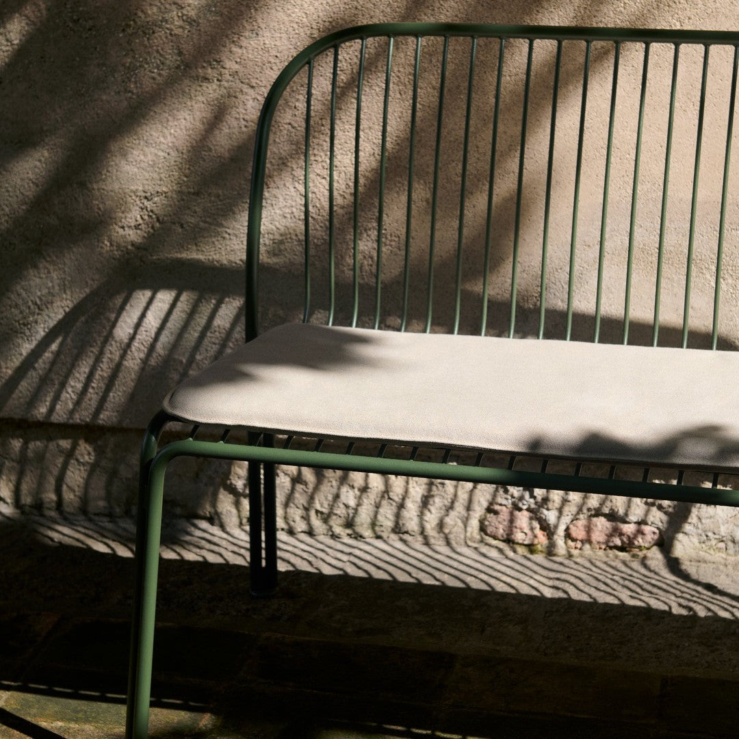 Green metal bench with a light beige cushion, featuring sleek vertical lines and subtle shadows.