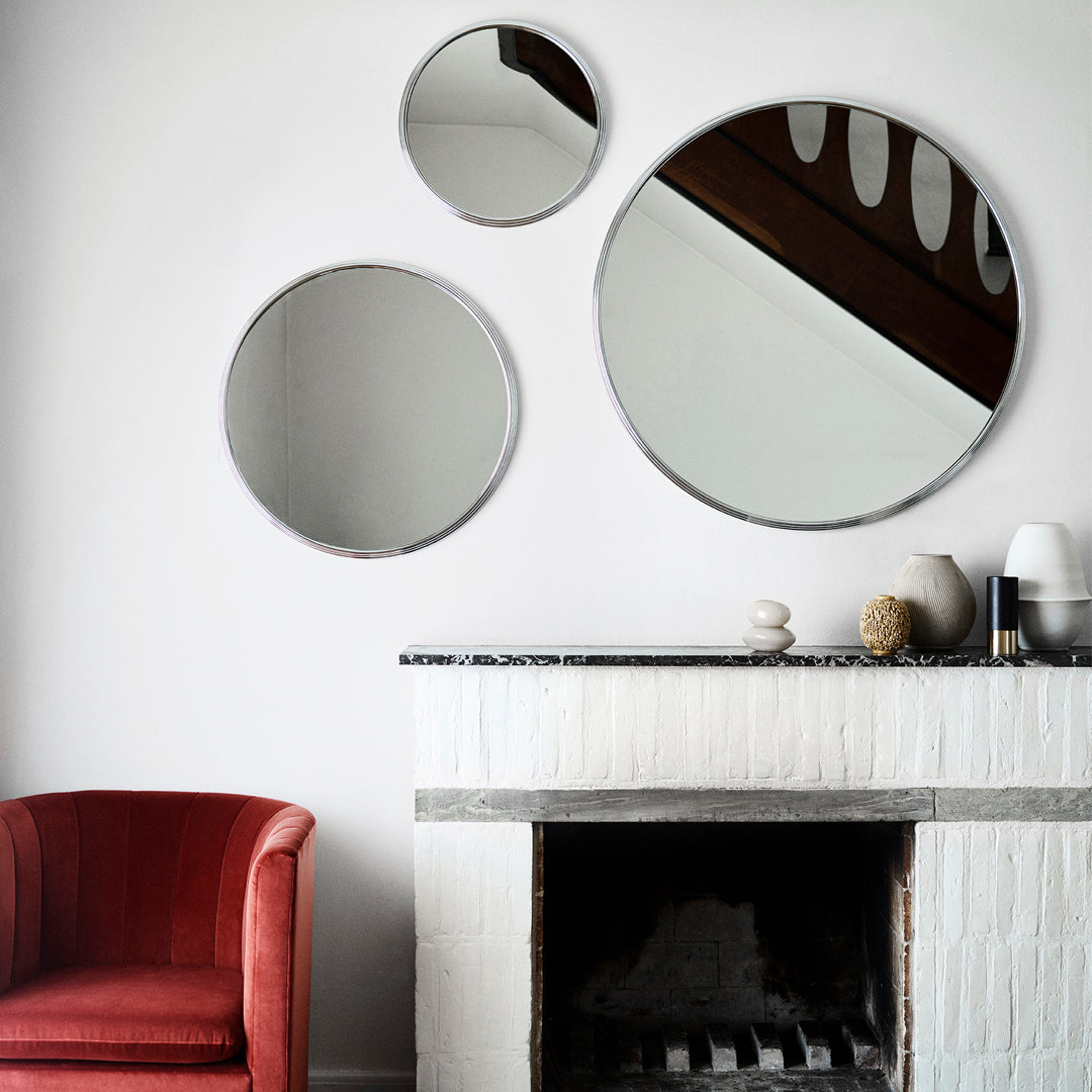 "Three circular mirrors in black and silver reflect a minimalist setting with a red armchair and white fireplace."