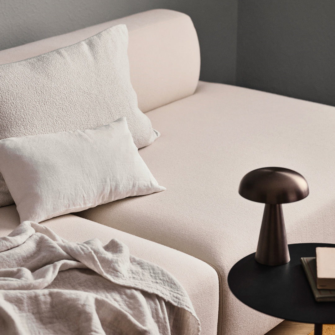 Light pink sofa with textured cushions, contrasting neutral throw blanket, and a sleek dark metal lamp on a round table.
