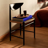 Black chair with a natural wood seat, featuring a blue book and brown pottery on top, against a warm background.
