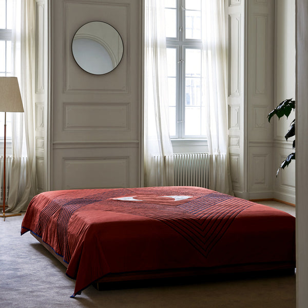 Red geometric patterned bedspread on a minimalist bed in a bright, airy room with white walls and natural light.