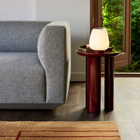 Gray sofa with rounded arm, red accent table, and soft glowing lamp, set against a natural backdrop and textured rug.