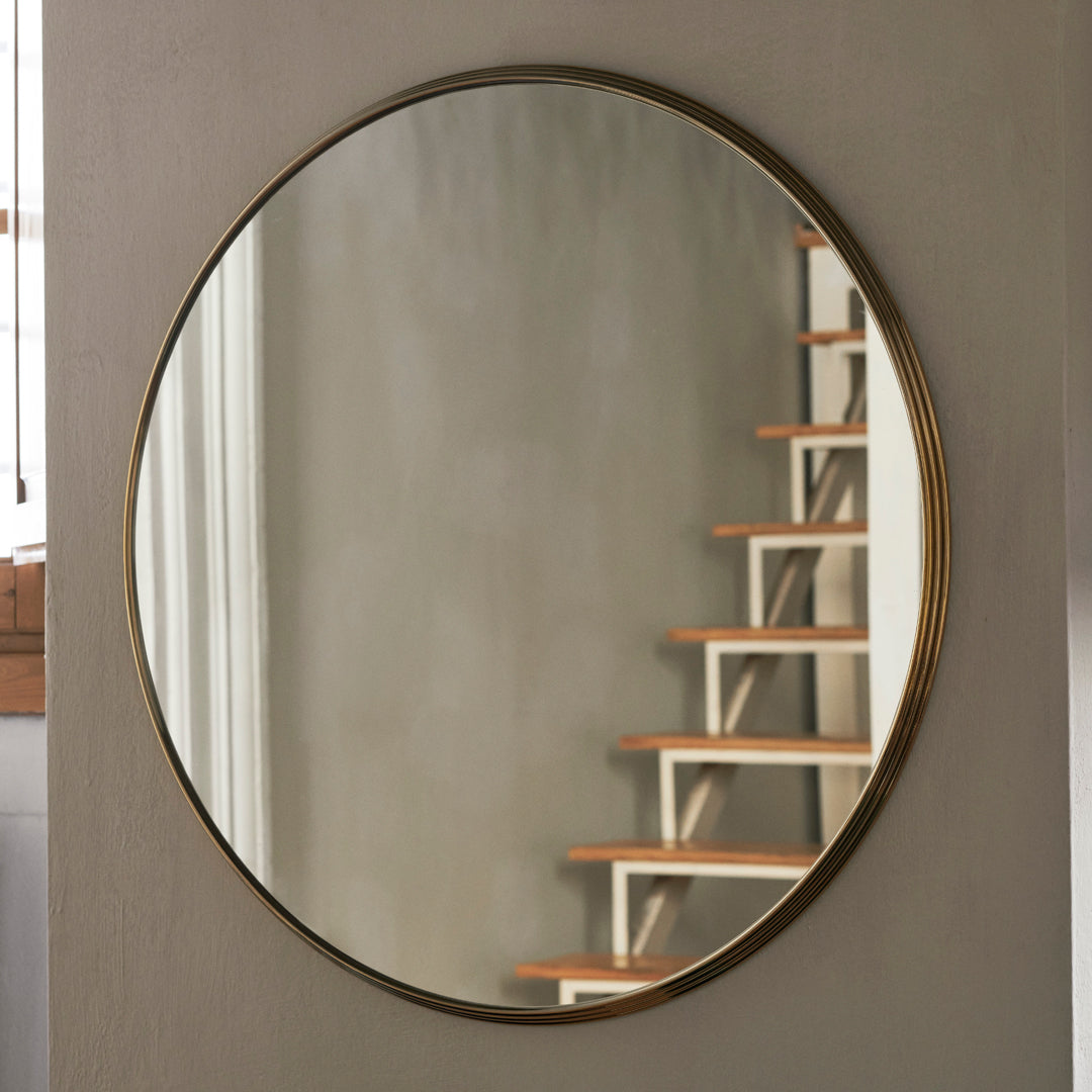 Round mirror with a sleek gold frame, reflecting a neutral wall and wooden staircase, adding elegance to any space.