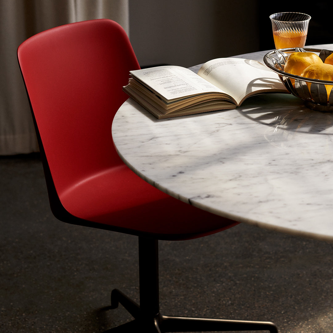 Red chair beside a round marble table with a casual book and a bowl of fruit, showcasing a sleek, modern design.