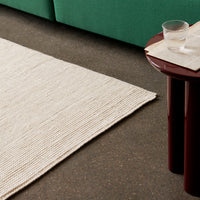 Beige textured rug beside a dark brown side table, with a green sofa in the background. Minimalist color palette.