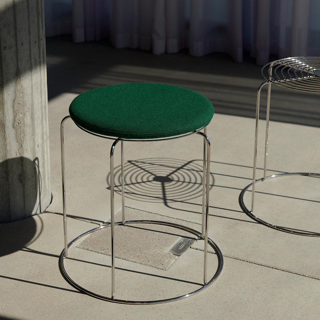 Green upholstered stool with a circular seat and sleek chrome frame, placed on a neutral floor with soft shadows.