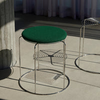 Green upholstered stool with a circular seat and sleek chrome frame, placed on a neutral floor with soft shadows.