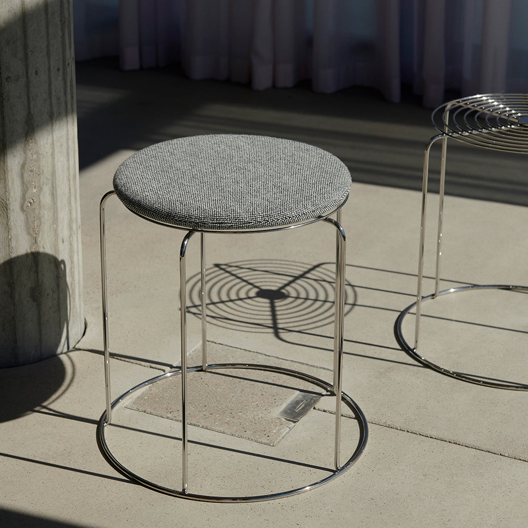 Gray upholstered stool with sleek chrome base, featuring a minimalist design and modern patterns. Perfect for contemporary decor.