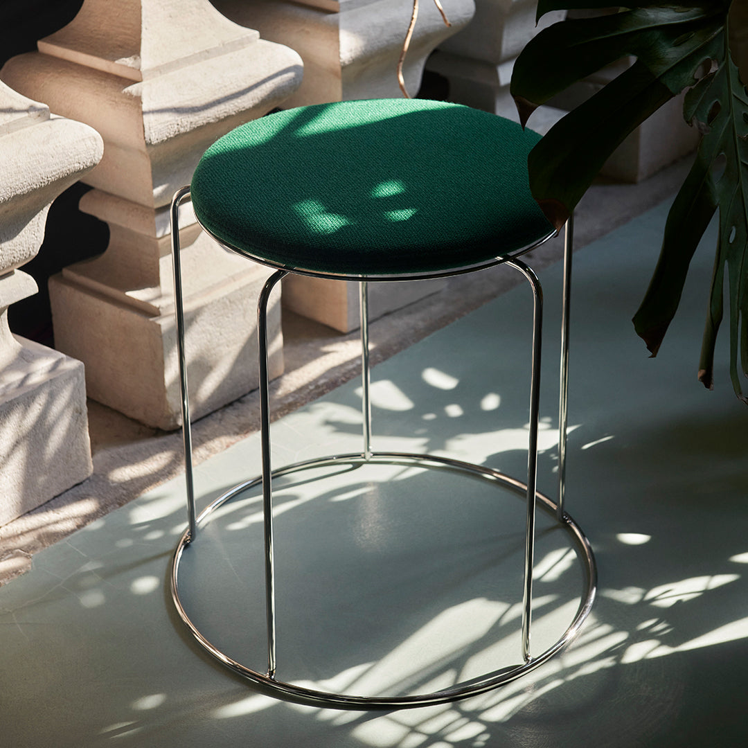Green upholstered stool with a sleek chrome base, featuring soft shadows and light patterns on the floor.