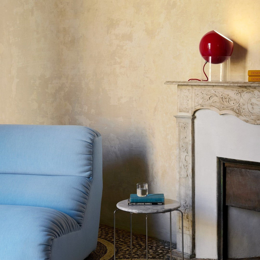 Light blue sofa with subtle texture, paired with a glossy red lamp and neutral beige walls. Elegant patterns below.