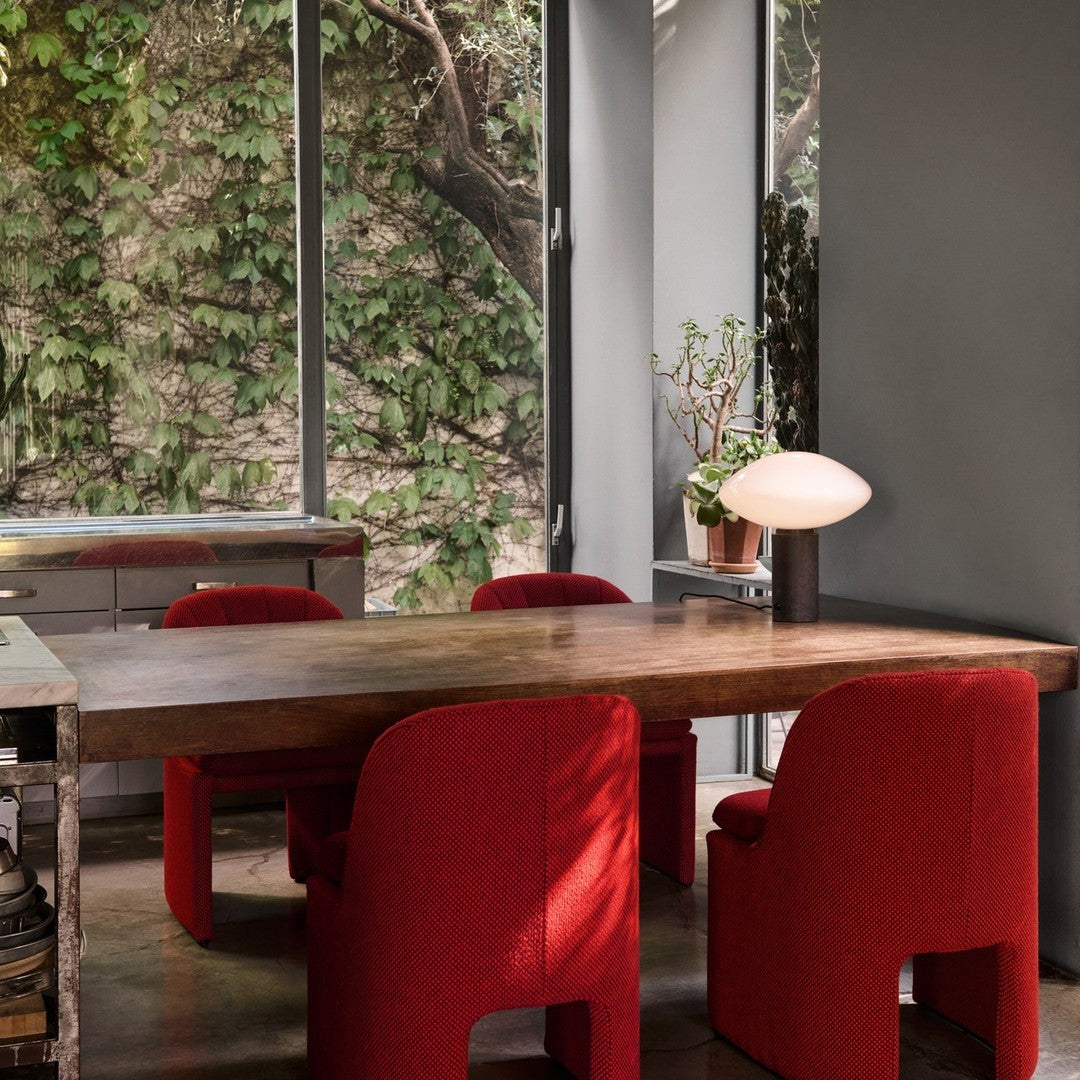 Red textured chairs surrounding a wooden table in a modern setting with green foliage and gray walls.