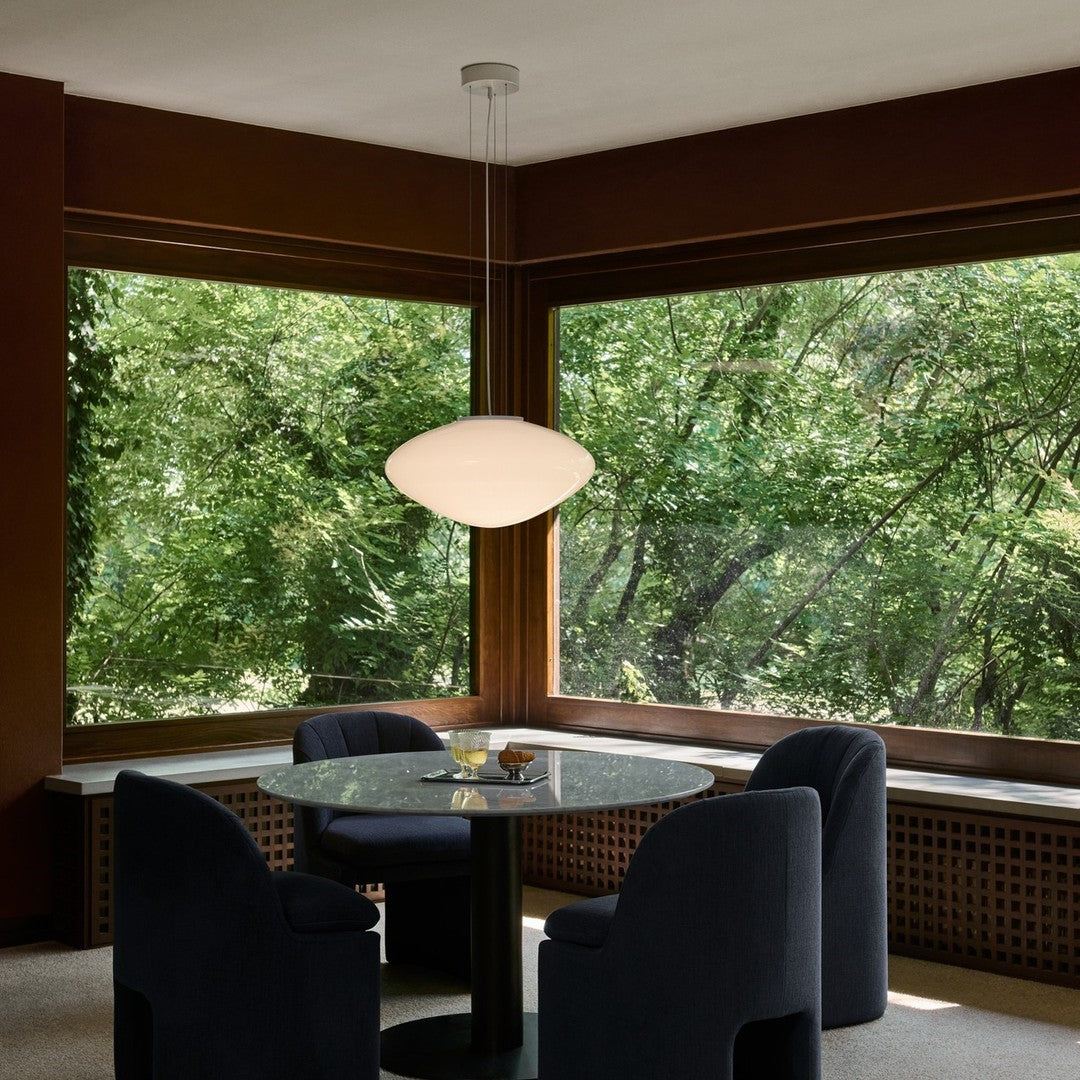 Modern pendant lamp, glossy white finish, black base, set against rich brown wood and lush green outdoor backdrop.