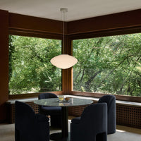 Modern pendant lamp, glossy white finish, black base, set against rich brown wood and lush green outdoor backdrop.
