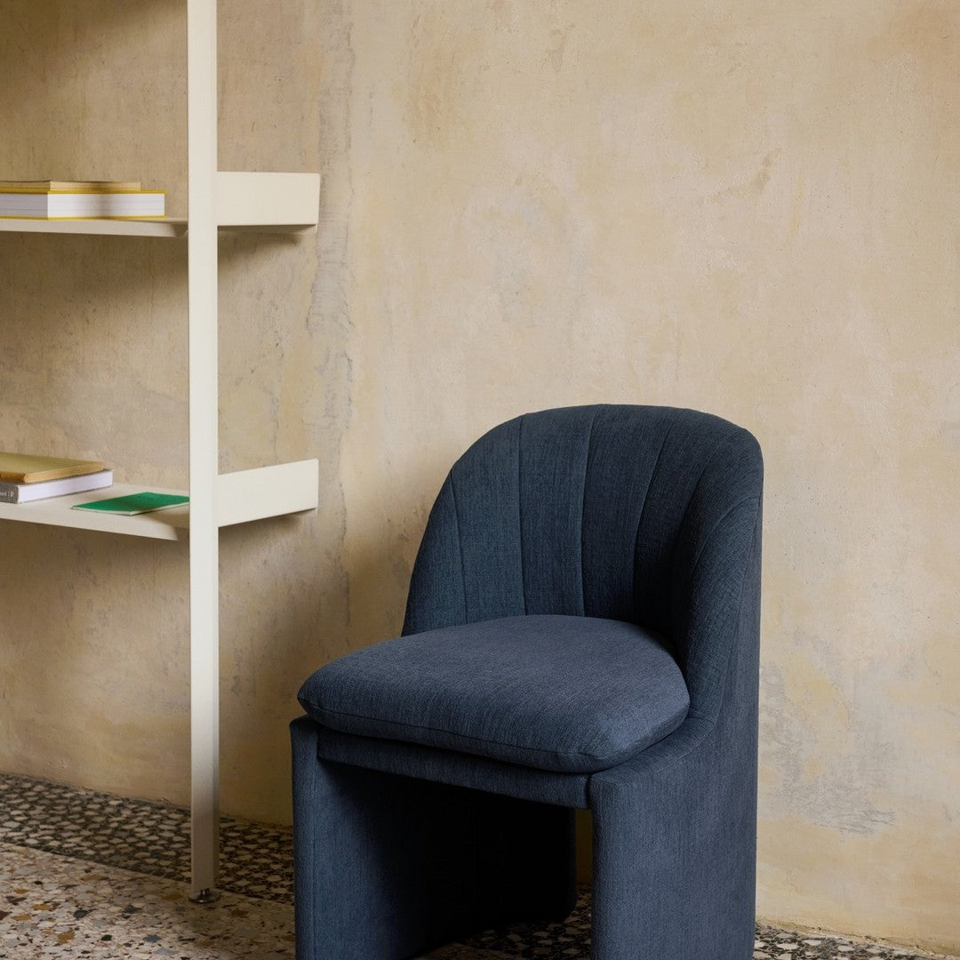 Dark blue upholstered chair with curved lines against a textured beige wall and light-colored shelving unit.