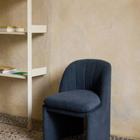 Dark blue upholstered chair with curved lines against a textured beige wall and light-colored shelving unit.
