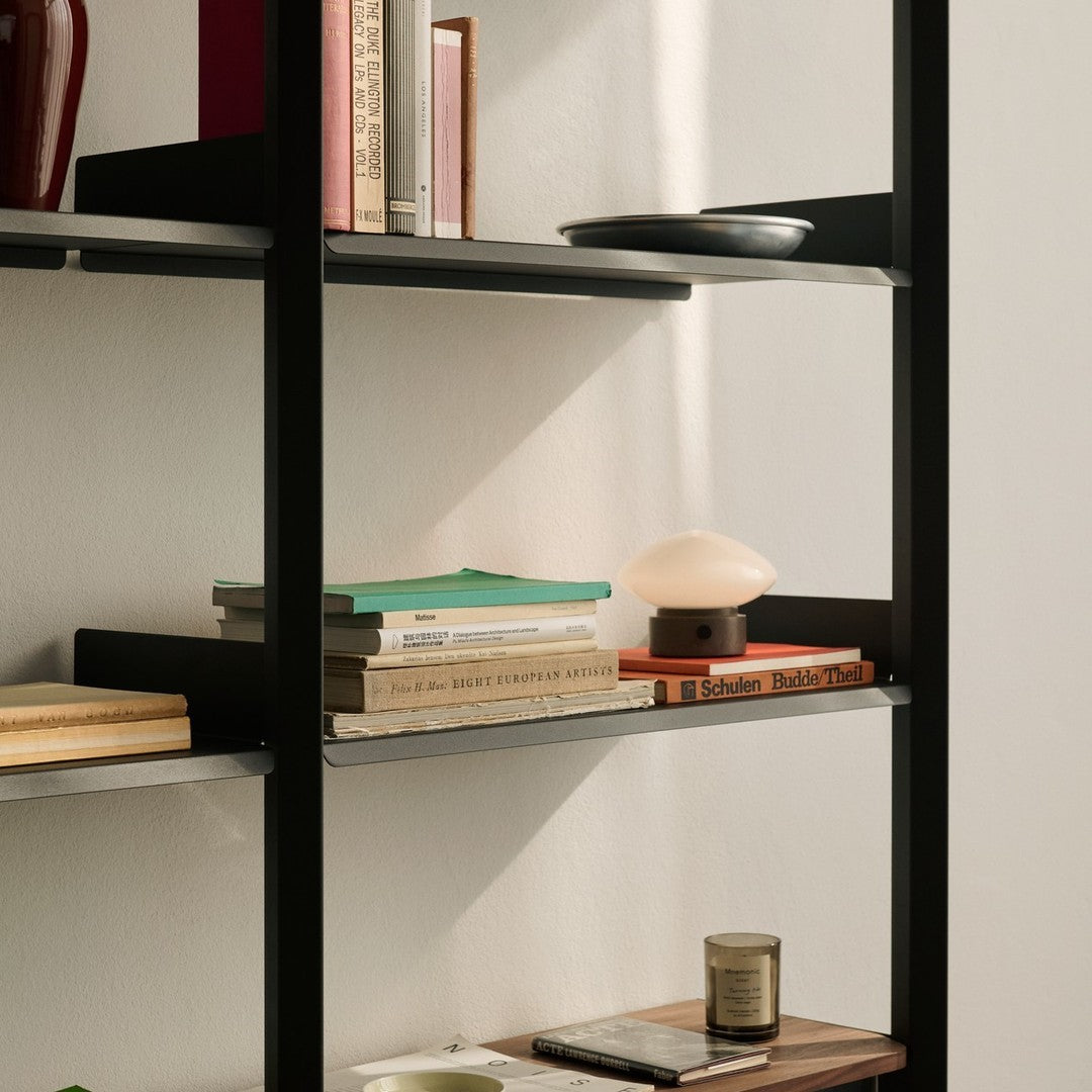Black shelving unit with colorful books, green and orange accents, and a soft-lit lamp against a neutral background.