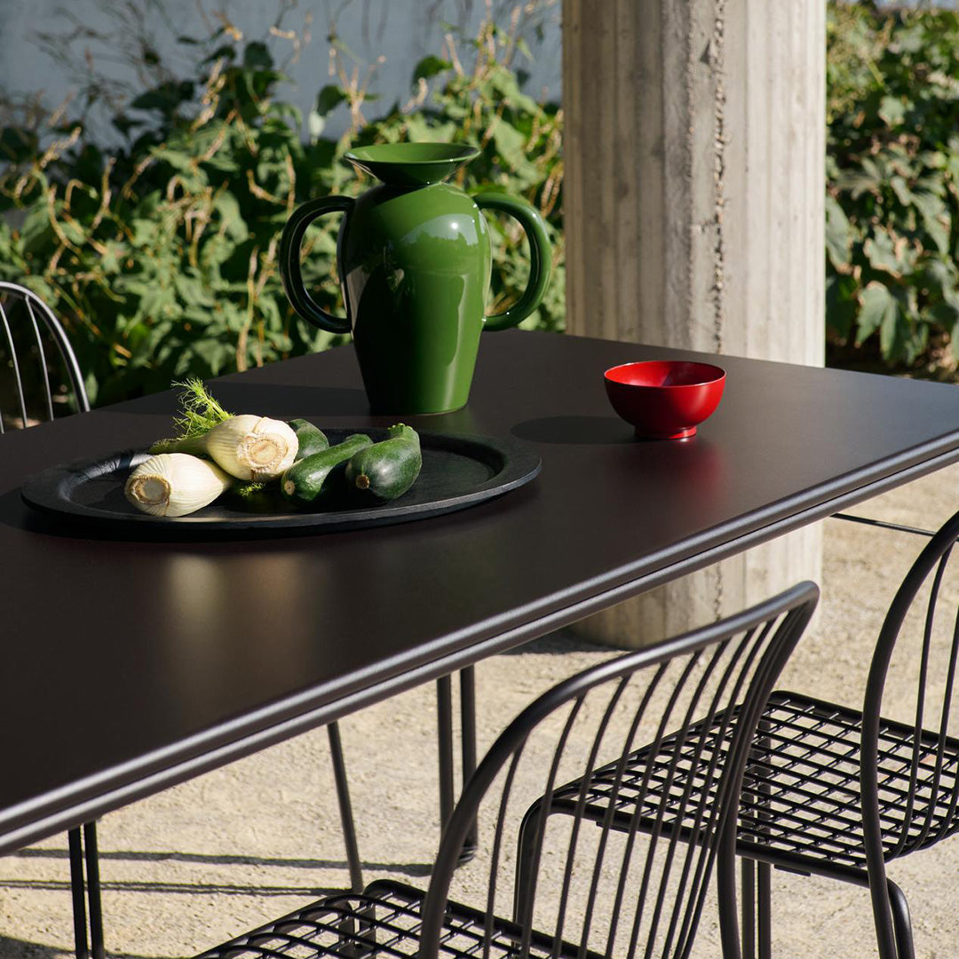 "Black tabletop with green ceramic pitcher and red bowl, surrounded by black wire chairs, set against lush greenery."