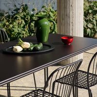 "Black tabletop with green ceramic pitcher and red bowl, surrounded by black wire chairs, set against lush greenery."