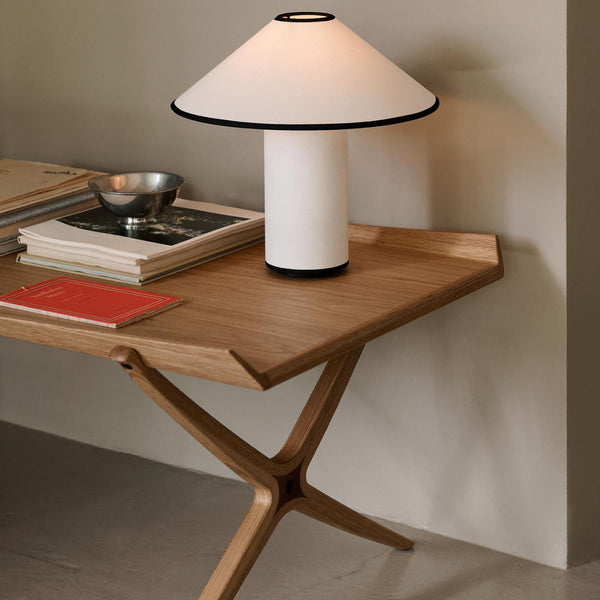 White lamp with black trim on a light wooden table, featuring stacked books and a metallic bowl. Soft neutral background.