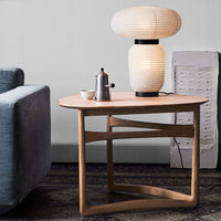 Light wood coffee table with an oval top and geometric legs, paired with a soft white lamp on a textured rug.