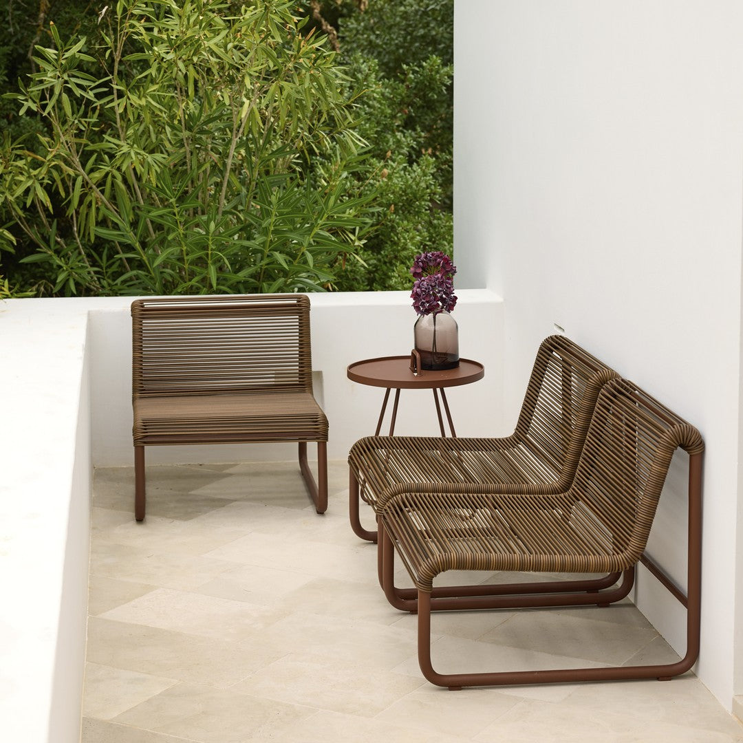 Brown woven patio chairs with a matching round table, set against a lush green backdrop and neutral flooring.
