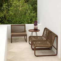 Brown woven patio chairs with a matching round table, set against a lush green backdrop and neutral flooring.