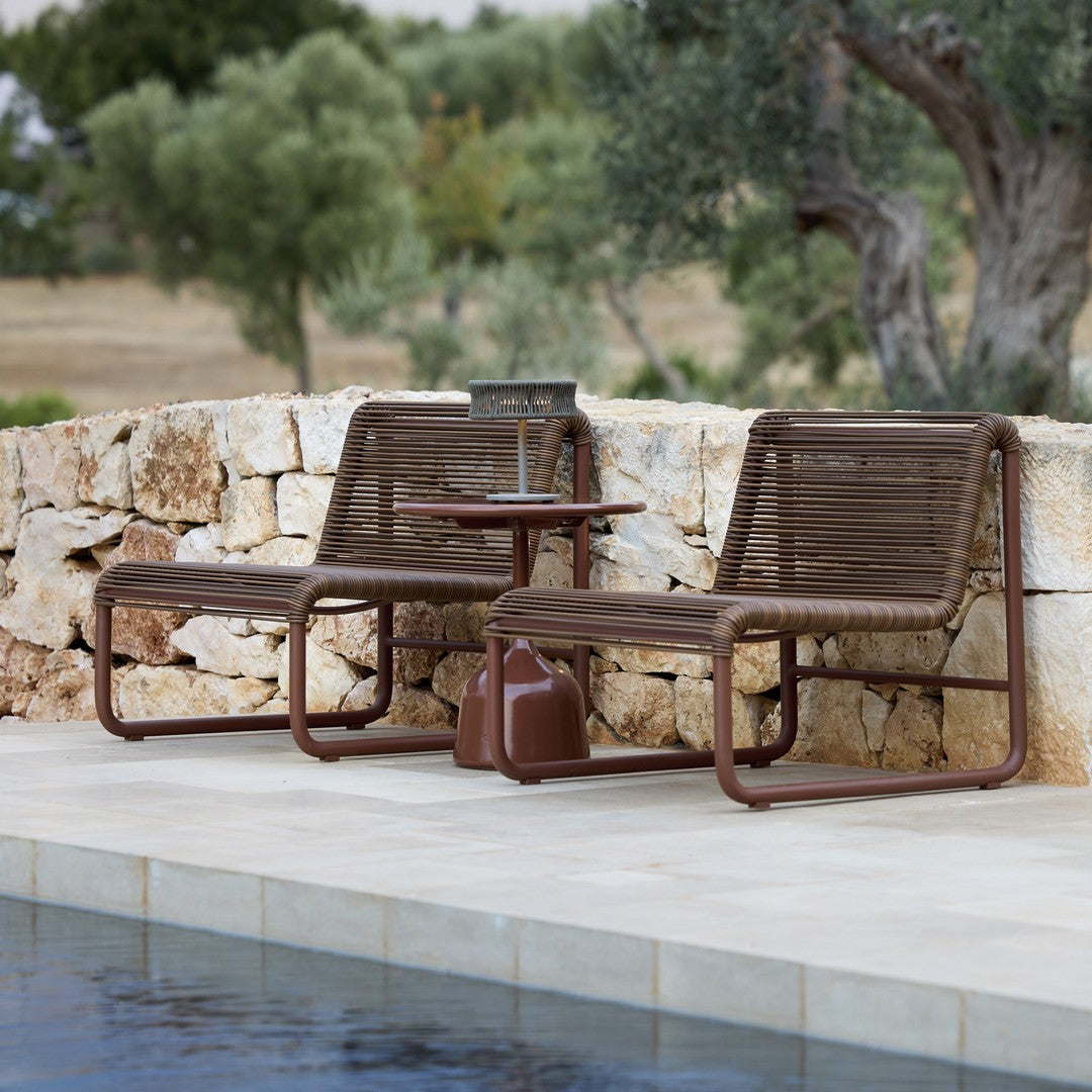 Brown metal frame lounge chairs with woven detailing, paired with a matching side table by a stone wall and pool.
