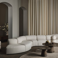 Curved beige sofa with soft texture, paired with dark wooden tables and light cream curtains in a modern setting.